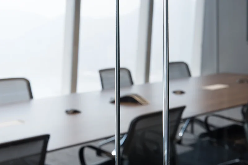 Minimalist office meeting room with a long white conference table, black chairs, and glass partitions.