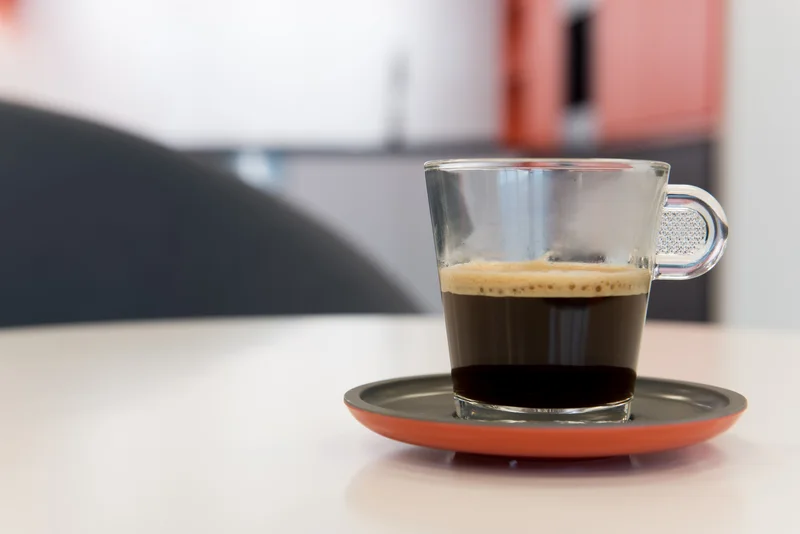 A glass cup of espresso on a dark saucer with a coral rim, set on a light table against a softly blurred modern interior.