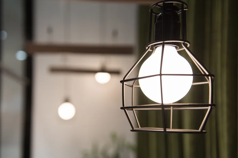 Close-up of a glowing spherical pendant light in a black geometric metal cage, with blurred lights and green curtains in the background.