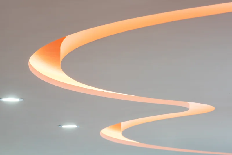 Abstract view of a sculptural curved ceiling light glowing warm orange against a pale gray office ceiling.