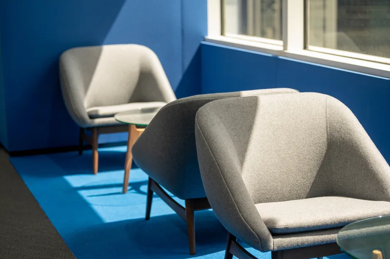 Contemporary lounge with gray upholstered armchairs, a glass table, vivid blue walls and carpet, and strong sunlight through windows.