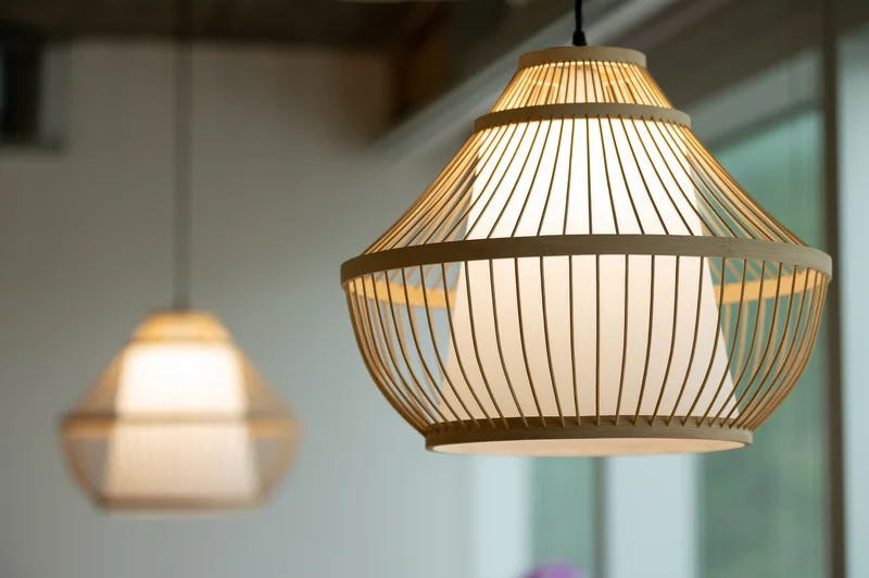 Close-up of a warm pendant light with a curved wooden slat shade, with a second blurred lamp in a modern interior.