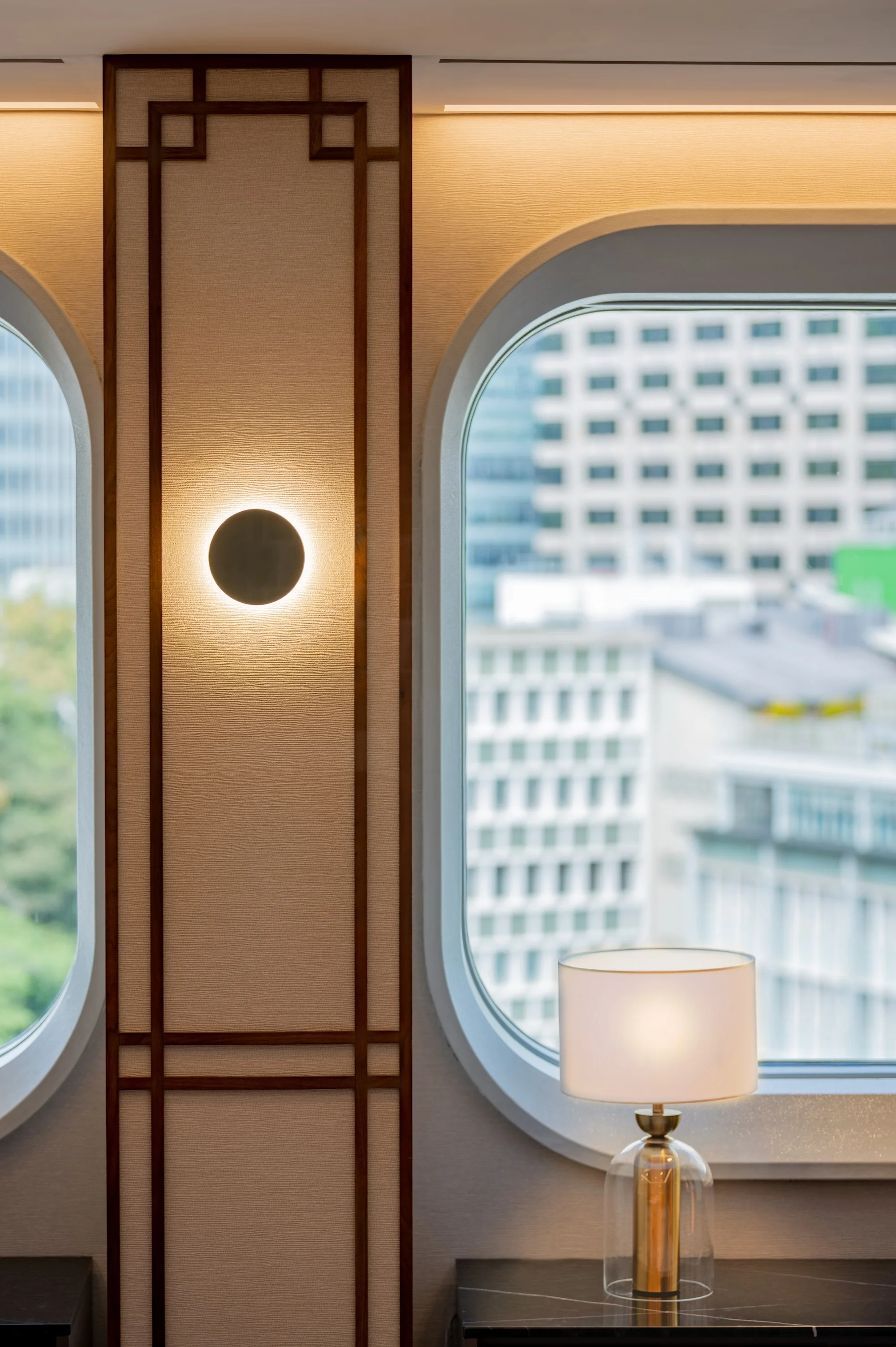 Wall panel with geometric wood trim in a traditional Asian lattice pattern, a glowing circular sconce, and oval windows framing a city view