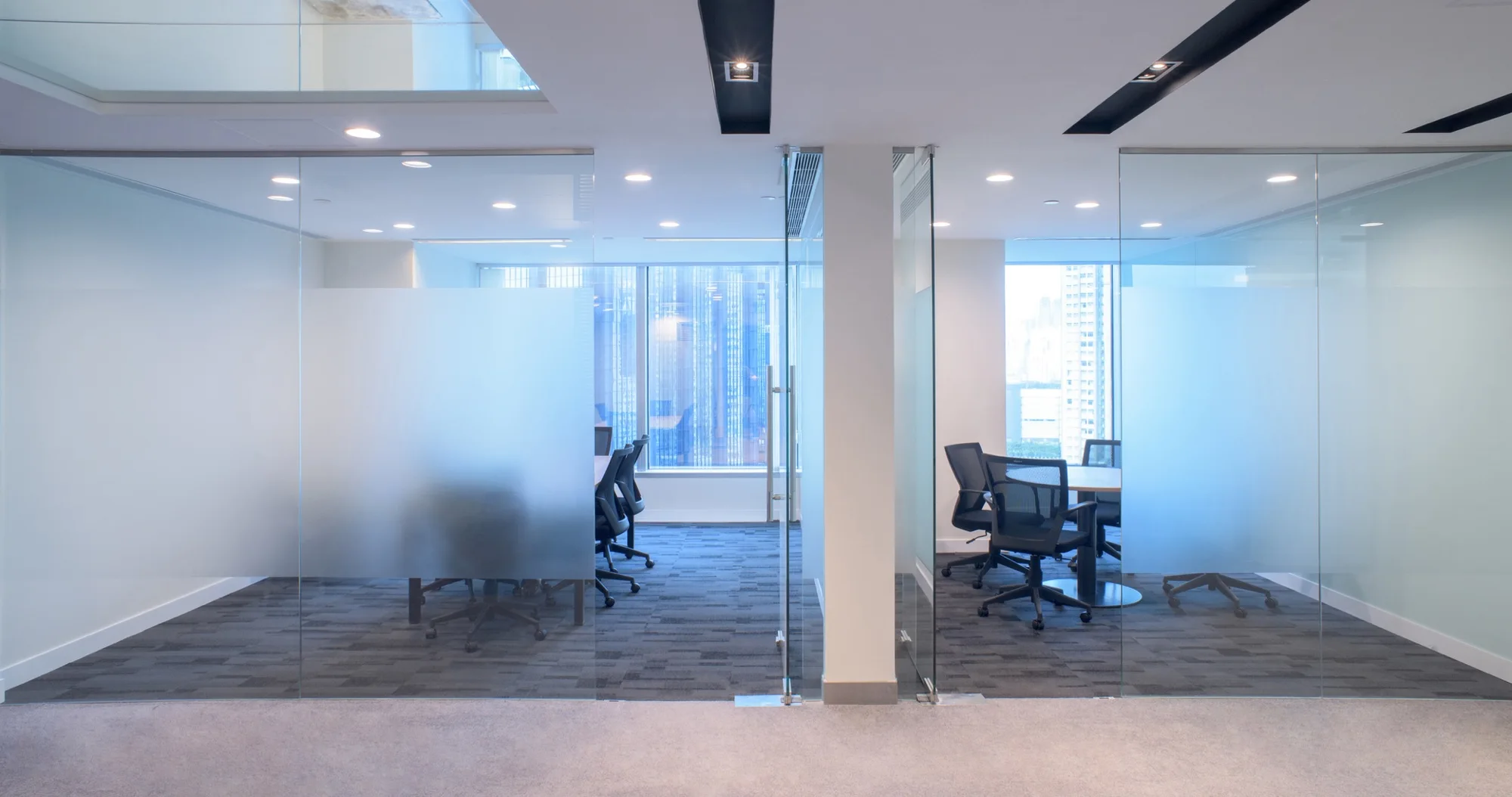 Glass-walled office meeting rooms with desks, black chairs, blue-gray carpet, recessed lighting, and city views.
