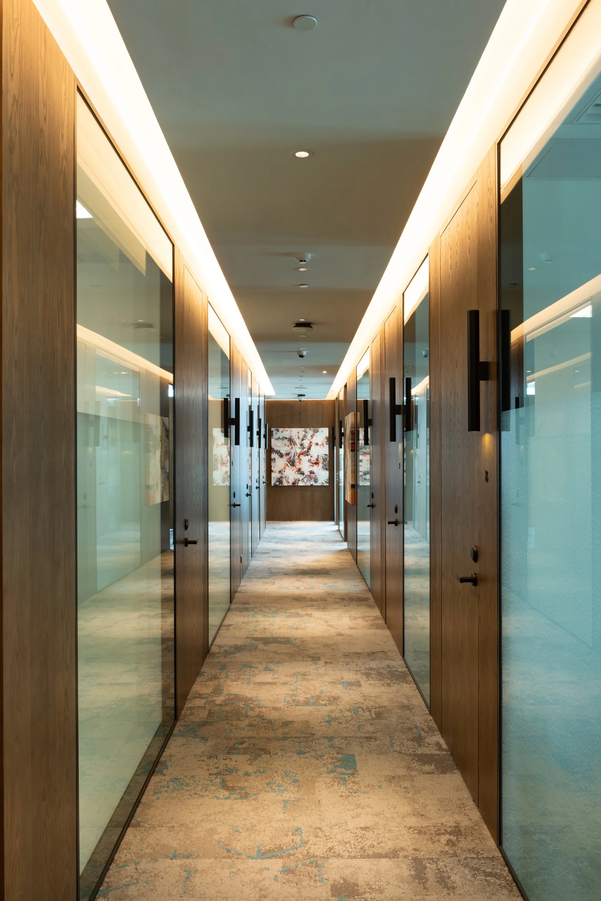 Modern office corridor with walnut paneling, glass-fronted rooms, patterned carpet, and illuminated ceiling coves.