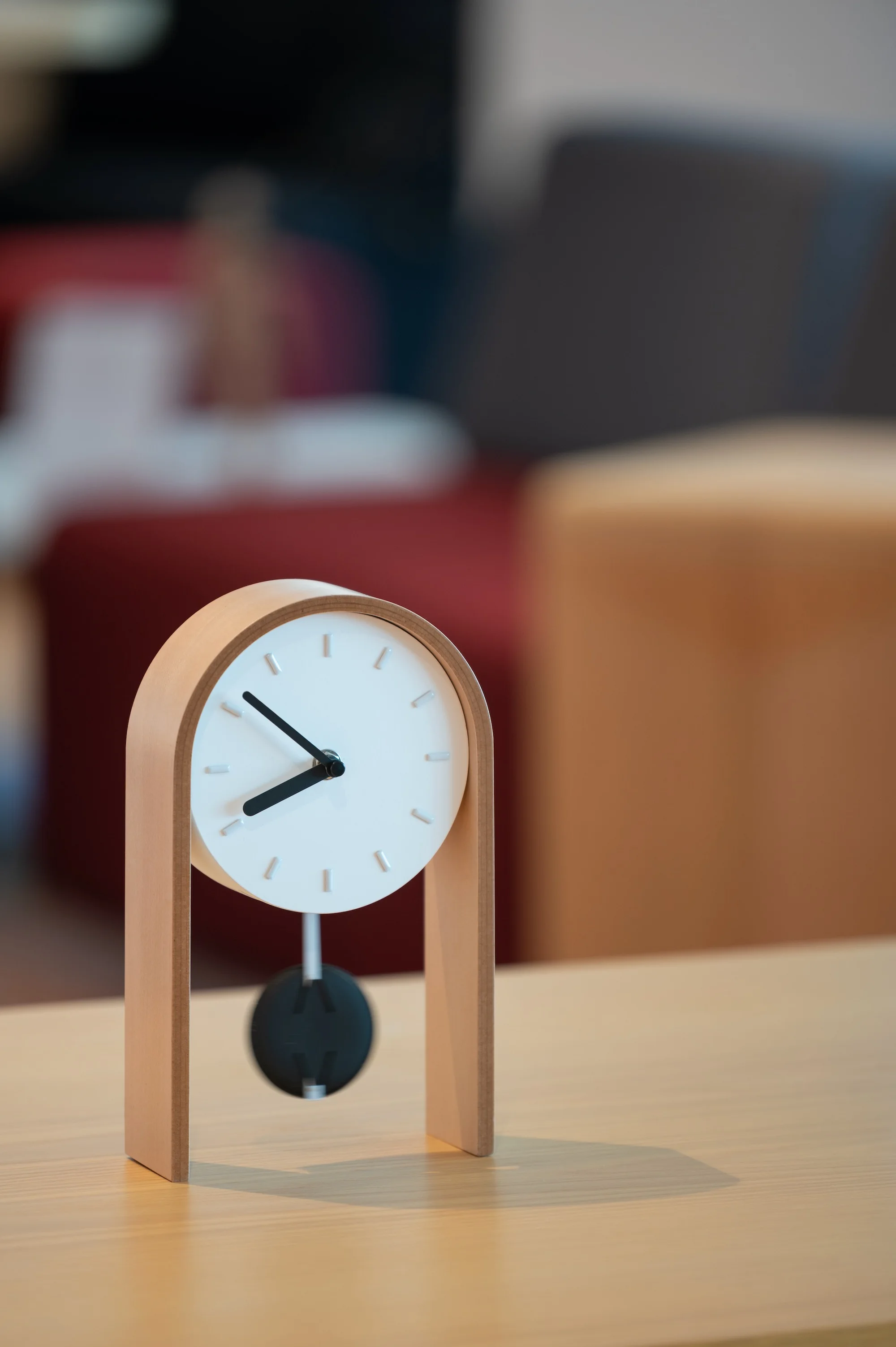 Minimal wooden mantel clock on a light wood desk with a softly blurred office interior in the background.