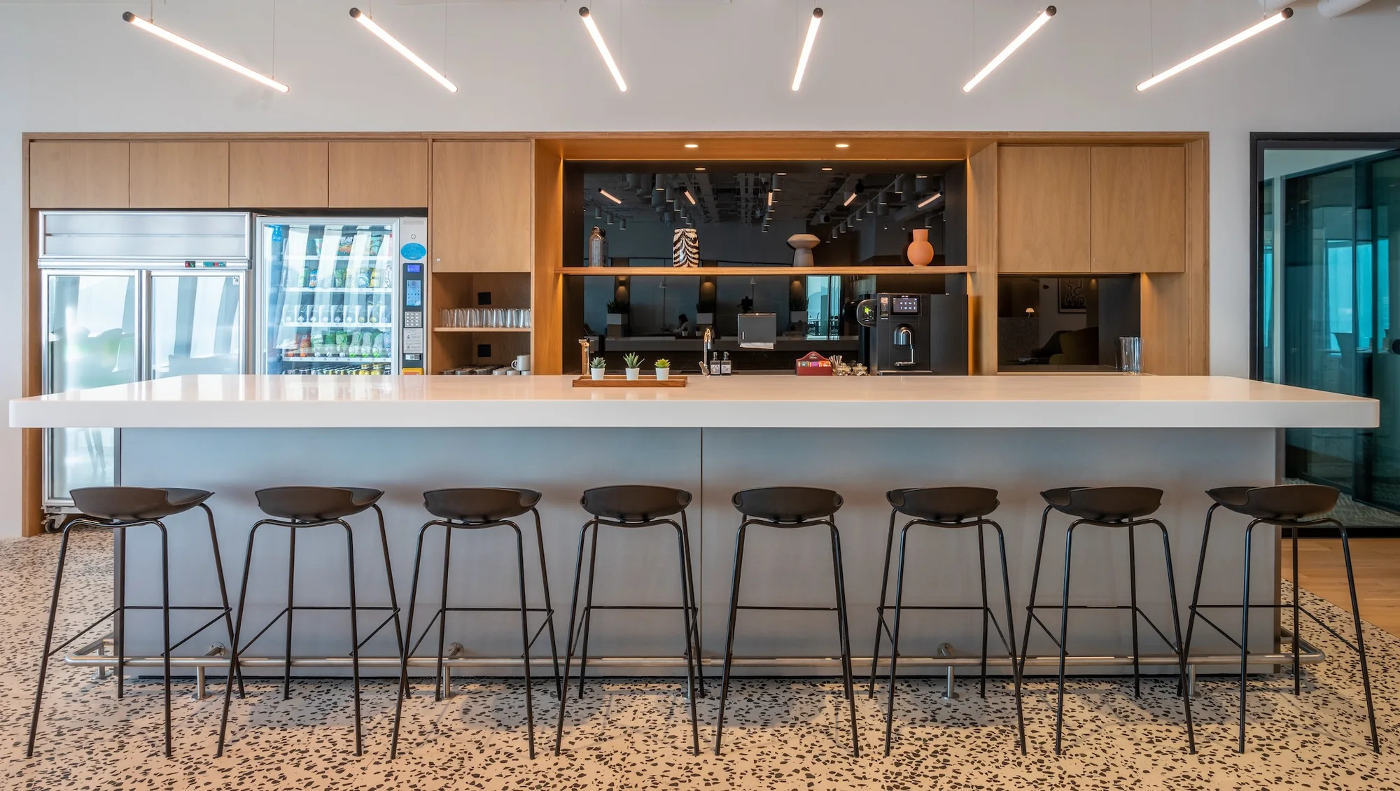 Premium serviced-office cafe with a long white counter, black bar stools, oak cabinetry, coffee station, and terrazzo flooring.