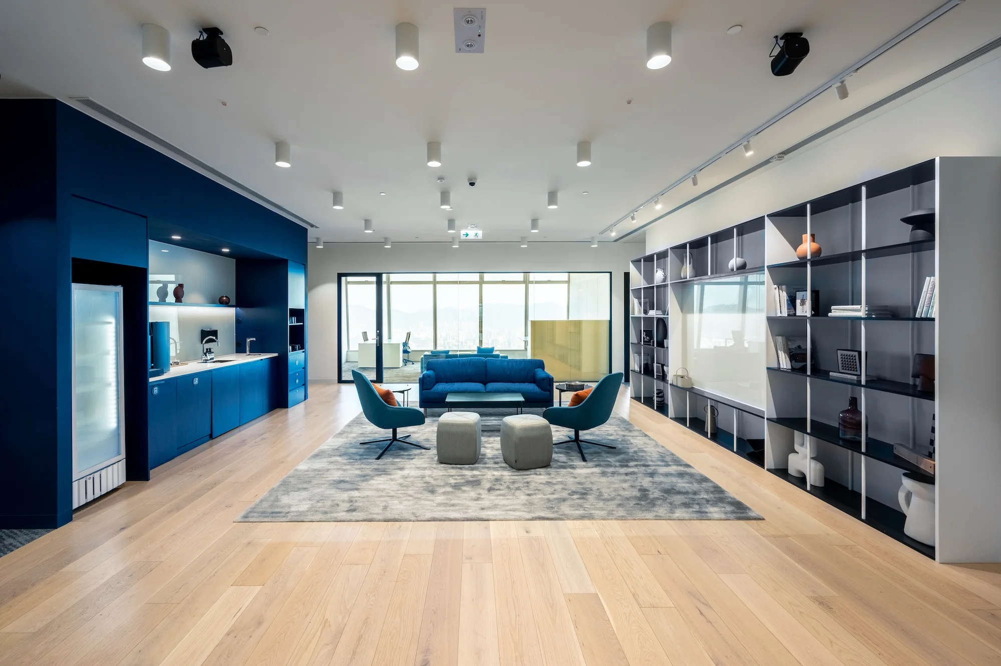 Premium serviced office lounge with blue seating, navy kitchenette, display shelving, pale wood flooring, and views from ICC Hong Kong.